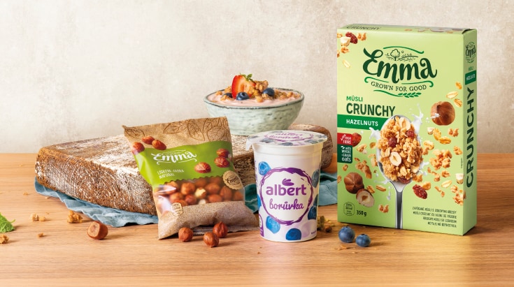 Snídaňové produkty zahrnující krabici müsli a sáček lískových oříšků značky Emma, doplněné kelímkem borůvkového jogurtu Albert a čerstvými borůvkami.