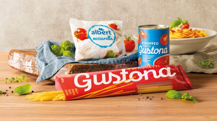 Suroviny pro přípravu italského jídla: špagety a krájená rajčata značky Gustona, mozzarella Albert, čerstvá rajčata a bazalka s hotovými těstovinami v pozadí.